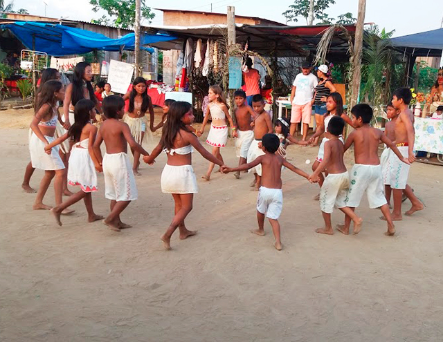 Em espaço aberto, a educadora indígena Vanda Witoto participa de uma roda de dança onde crianças estão de mãos dadas. Todos vestem roupas claras. Alguns estão com pinturas no rosto. Fim da descrição. 