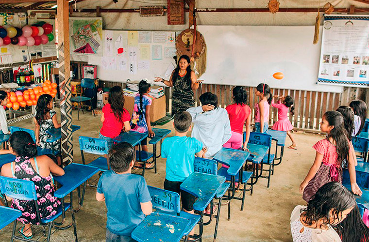 No CMEEI, Claudia Baré está de frente para várias crianças, que estão de pé ao lado de carteiras escolares. No lado esquerdo do espaço de aulas, há bexigas coloridas sobre mesa e na parede. Fim da descrição.
