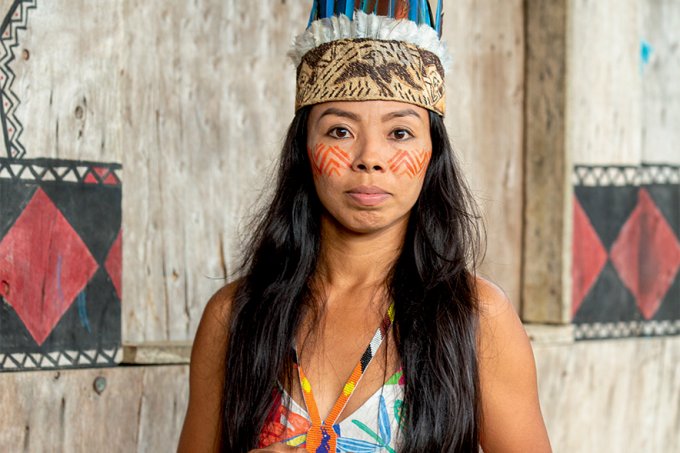 Vanda Witoto, em frente à estrutura de madeira, olha para a câmera. Ela usa cocar, colar colorido e pintura tradicional no rosto. Fim da descrição.