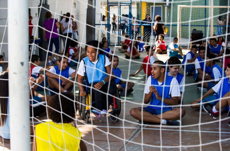 Estudantes realizam atividade em quadra esportiva. Os adolescentes estão sentados no chão. Uma garota em uma cadeira de rodas sorri no centro da cena.
