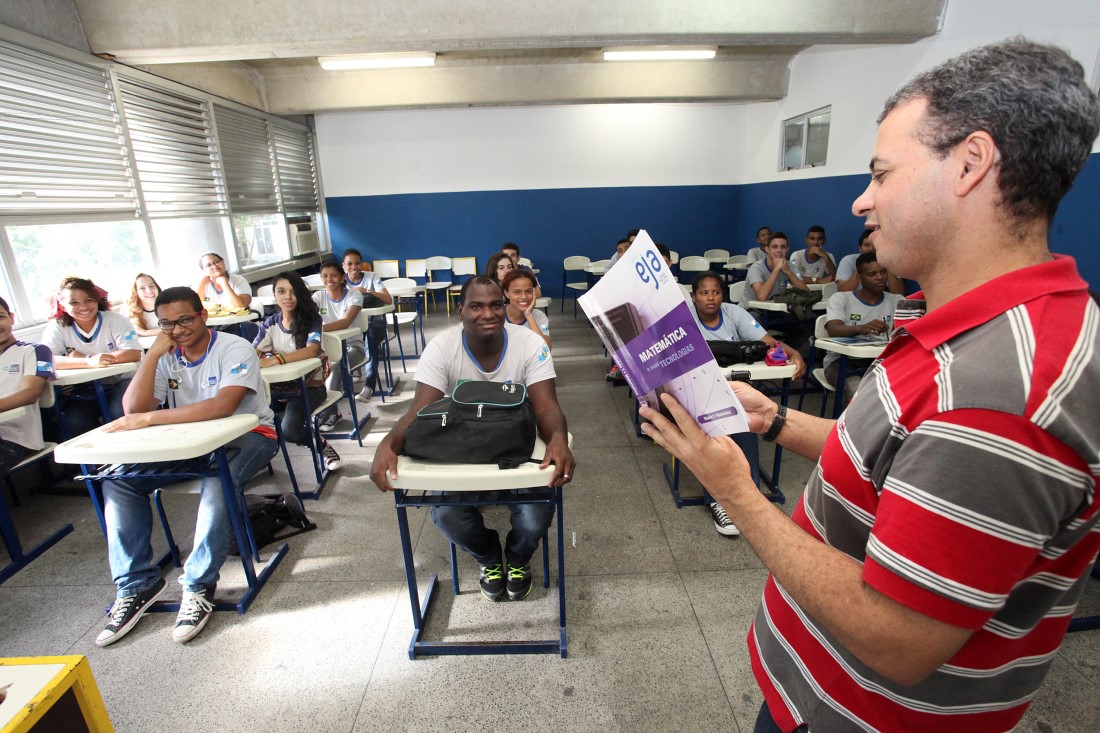 Em sala de aula, professor está em pé segurando um livro de matemática.  Ao fundo, estudantes uniformizados estão sentados. Fim da descrição.