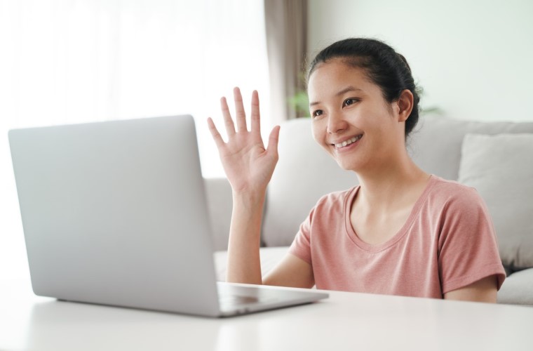 Em sala de estar, uma mulher branca asiática está sentada à frente do notebook e faz um sinal com a mão direita enquanto olha para a tela. Fim da descrição.