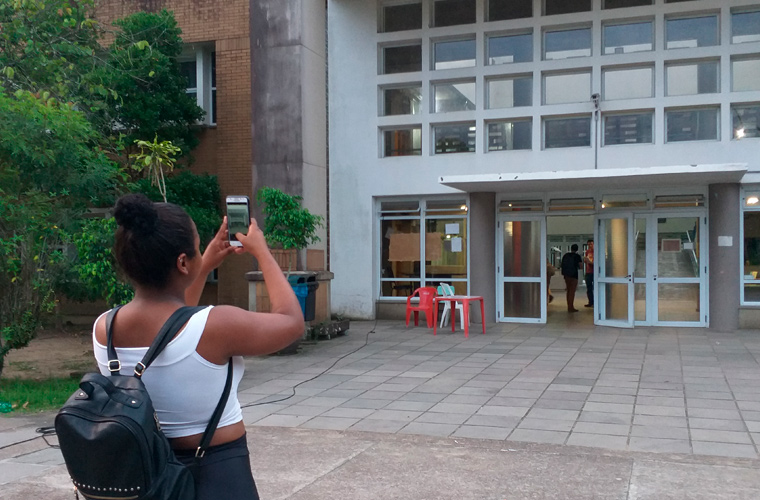 Em espaço externo, estudante negra com mochila nas costas tira foto de prédio da escola com o celular. Fim da descrição.
