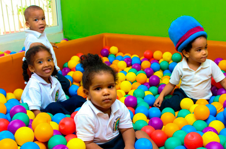 Quatro crianças negras, sendo três meninas e um menino, estão brincando dentro de uma piscina de bolinhas coloridas. Fim da descrição.