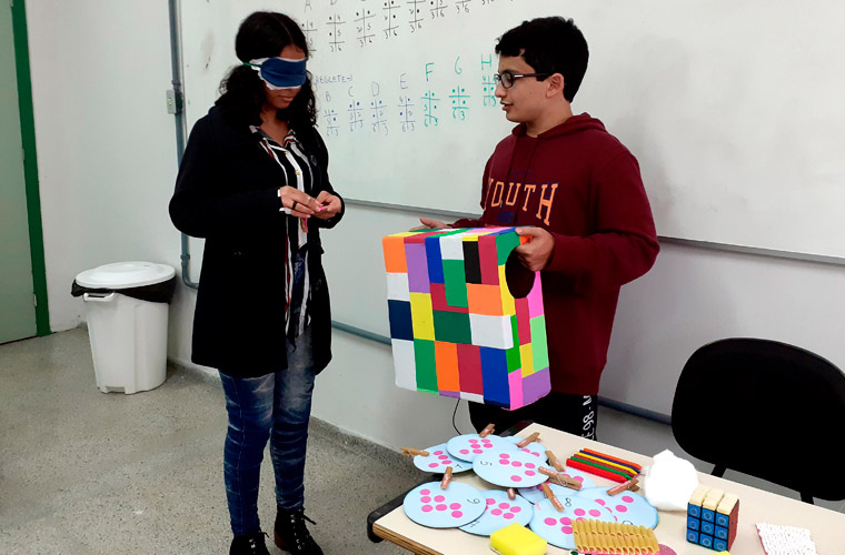 Em sala de aula, uma estudante do curso “Braille básico” está vendada enquanto um colega segura caixa colorida com entradas circulares nas laterais. Fim da descrição.