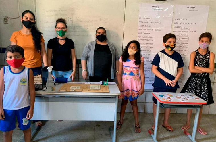 De pé em sala de aula, quatro estudantes, duas meninas e dois meninos, e três educadoras posam para foto, com quadro branco ao fundo. Todos usam máscara de proteção no rosto. Fim da descrição.