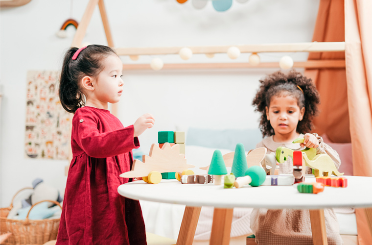 Duas meninas, uma asiática e uma negra, estão perto de uma mesa redonda com brinquedos de madeira. Fim da descrição.