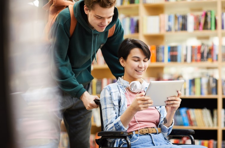 Em uma biblioteca, dois estudantes, uma mulher e um homem brancos, observam um tablet. A estudante está em cadeira de rodas. Fim da descrição. 