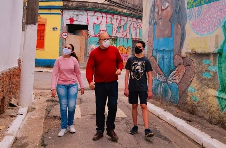 Educador e dois estudantes, sendo uma menina e um menino, estão caminhando por uma rua do bairro do Glicério, em São Paulo, com alguns muros grafitados. Todos usam máscara. Fim da descrição.