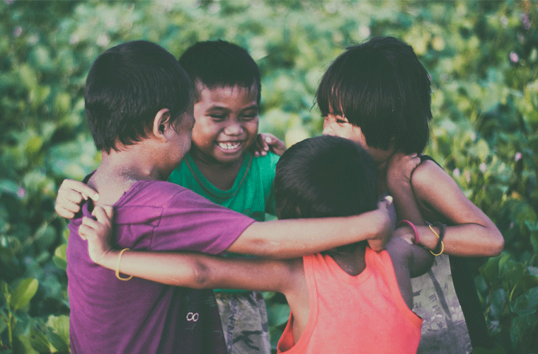Quatro crianças se abraçam em roda e sorriem. Ao fundo, muro de plantas.
