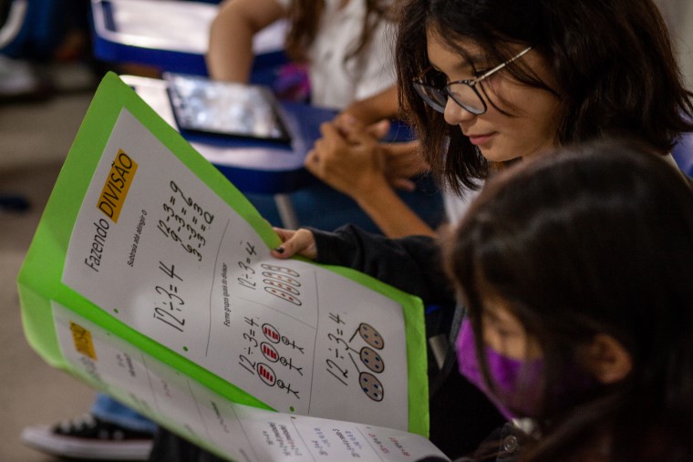 Estudantes em sala de aula. Em destaque, duas meninas, uma com máscara e outra sem, olham para o “caderno de cartazes”, que mostra maneiras de como solucionar operações matemáticas básicas. Fim da descrição.