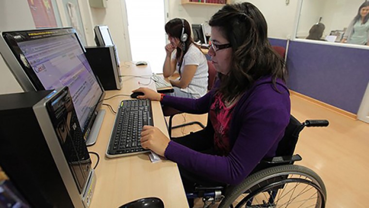 Duas mulheres estão em uma sala enquanto utilizam um computador. Uma das mulheres está em cadeira de rodas. Fim da descrição.
