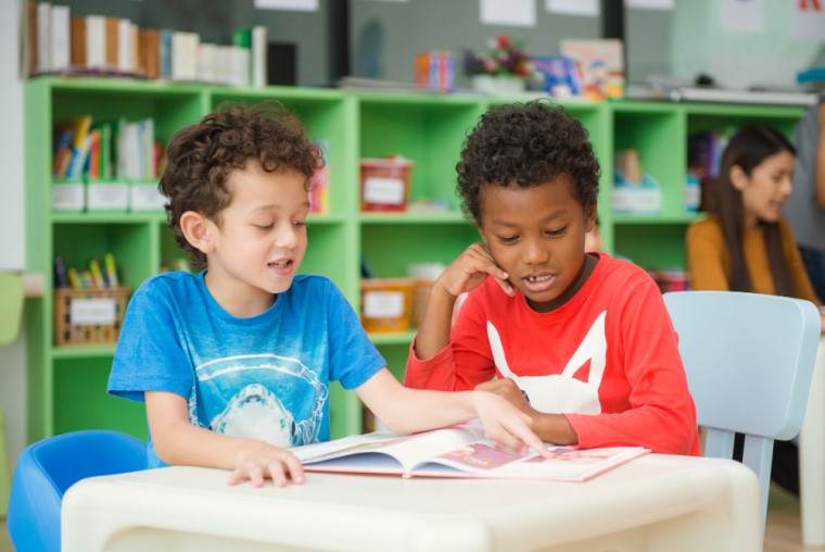 Em sala de aula, dois meninos, um branco e um negro, estão sentados observando um livro. Fim da descrição.