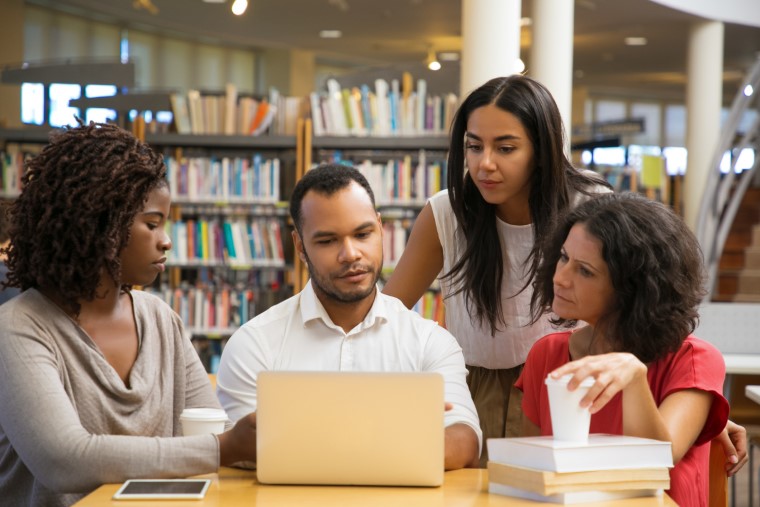 Em biblioteca, um homem negro, duas mulheres brancas e uma negra estão reunidos observando tela de notebook. Fim da descrição.