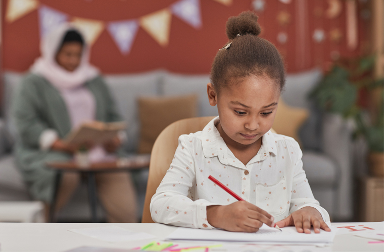 Em sala de aula, menina negra escreve em papel com lápis. Fim da descrição.