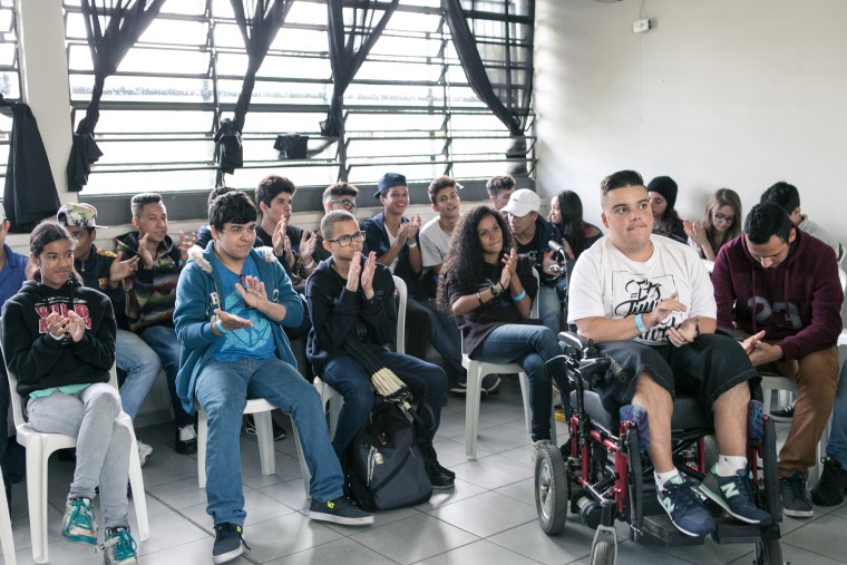 Em sala de aula, alunas e alunos adolescentes estão sentados enquanto batem palmas. Um dos alunos está em cadeira de rodas. Fim da descrição.