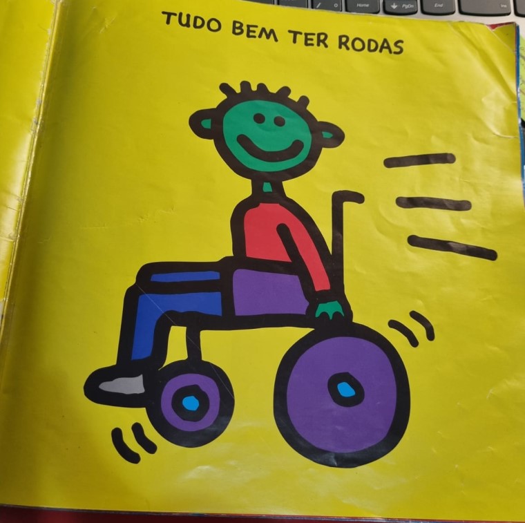 Livro aberto em página amarela, com ilustração de pessoa em cadeira de rodas. Acima, texto “tudo bem ter rodas”. Fim da descrição.