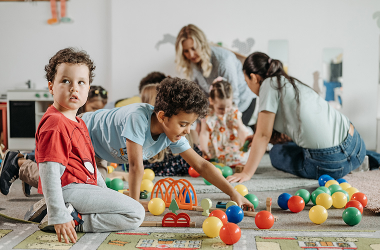 Em sala de aula de ensino infantil, dois meninos brincam com bolas coloridas e outros objetos sentados no chão. Ao fundo, há outras crianças e duas educadoras brincando. Fim da descrição.