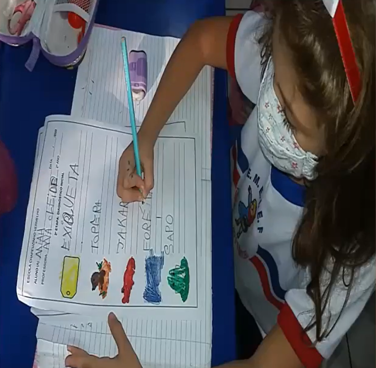 Menina uniformizada e de máscara escreve palavras a lápis em folha de papel com desenhos coloridos. Fim da descrição. 