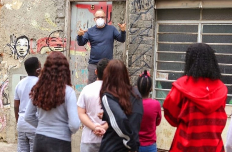 Educador realiza explicação a seis estudantes em uma rua do bairro da escola durante o projeto “Aula Pública”. Fim da descrição.