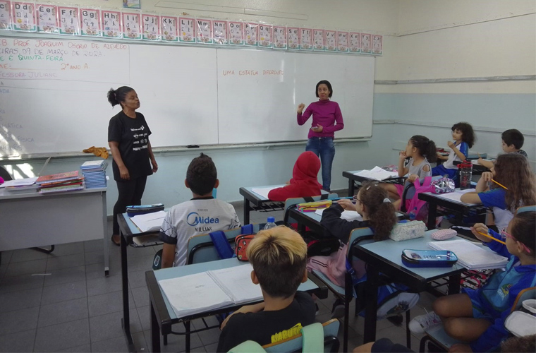 Em sala de aula, professora regente e intérprete de Libras ensinam um sinal em Língua Brasileira de Sinais para alunas e alunos que estão sentados nas carteiras. Fim da descrição.