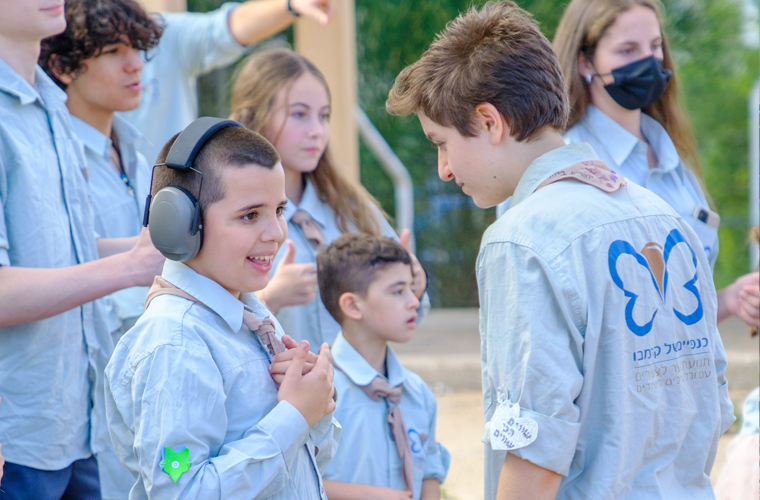 Jovens brancos com camisas da Krembo Wings estão em espaço aberto. Um menino com headsets fala com outro, que está à sua frente. Fim da descrição.