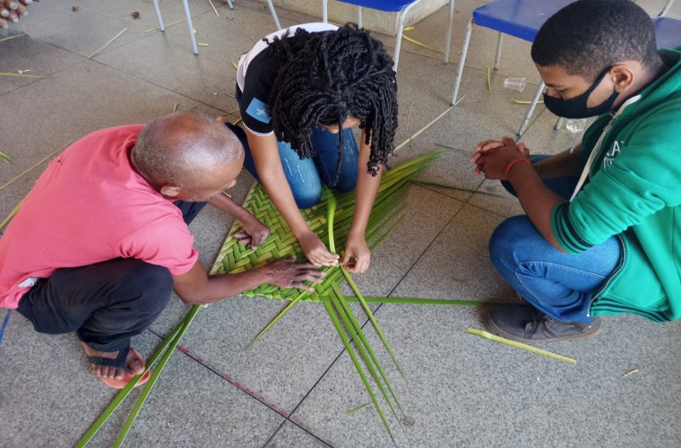 Uma estudante e um morador da comunidade estão agachados enquanto fazem artesanato ecológico com fibras das palmeiras oriundas da vegetação nativa. Ao lado, outro estudante os observa. Fim da descrição.
