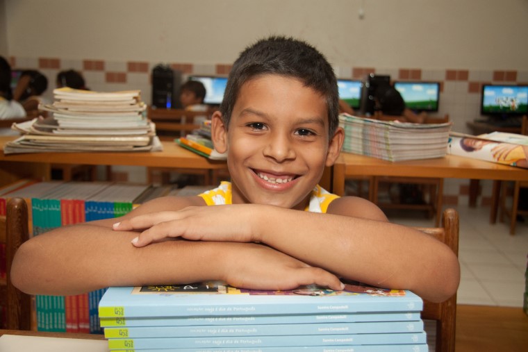 Em sala de aula, menino sorri enquanto está debruçado em uma pilha de livros. Fim da descrição.