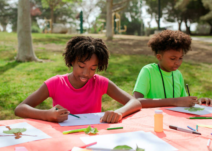 Imagem ilustrativa de artigo sobre dislexia. Na foto, uma menina e um menino negros estão ao ar livre e fazem desenho em sulfite. Fim da descrição.