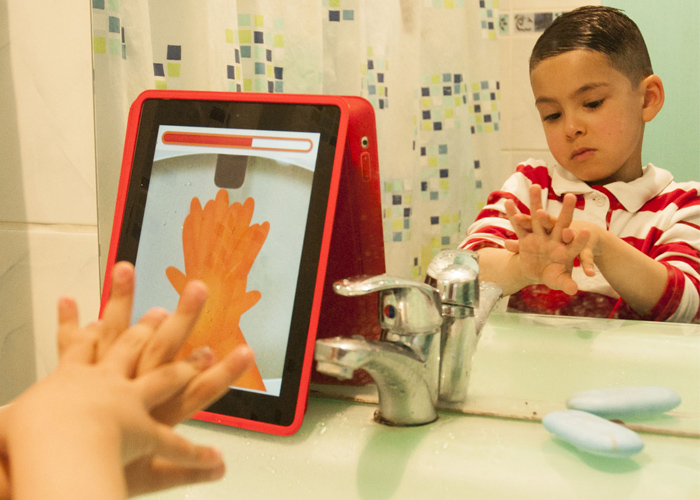 Em banheiro, menino branco está em frente a um espelho e copia gestos de lavar as mãos, mostrados em tablet que está em cima da pia, ao lado da torneira e de sabonete. Fim da descrição.