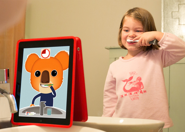 Em banheiro, menina branca está em frente a um espelho e copia gestos de escovar os dentes, mostrados em tablet que está em cima da pia, sendo feitos por um desenho de coala. Fim da descrição.