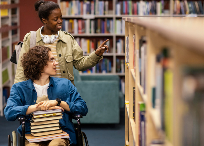 Uma jovem negra está em pé enquanto auxilia um jovem branco que está em cadeira de rodas com livros no colo. Ambos observam livros que estão na prateleira da biblioteca. Fim da descrição.