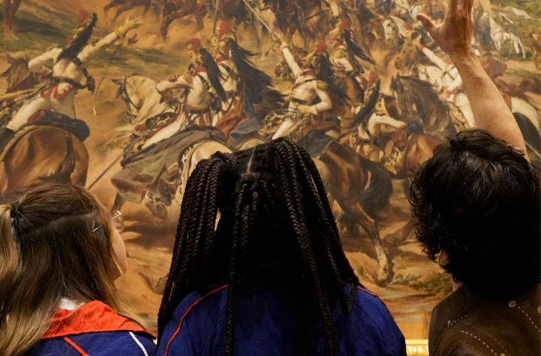 Três pessoas de costas diante da tela “Independência ou Morte”. Sobre a imagem está escrito: Encontro com professores no Museu do Ipiranga. Fim da descrição.