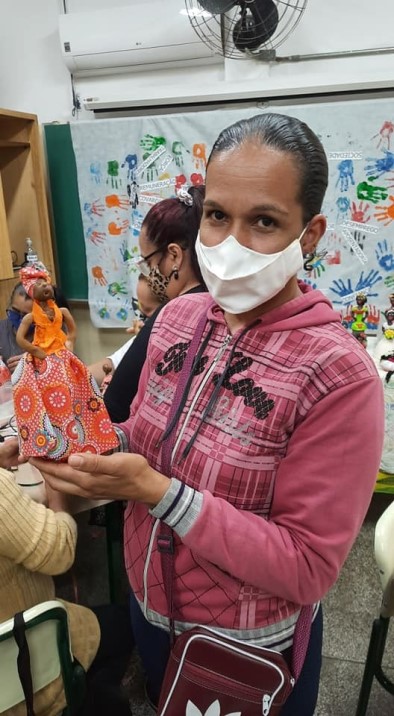 Uma mulher está em pé e segura uma boneca de pano confeccionada na própria escola. Ela usa máscara no rosto. Fim da descrição.