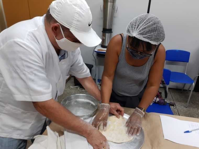 Um homem e uma mulher estudantes da EJA fazem massa de pizza. Ambos usam máscara no rosto e touca de proteção na cabeça. Fim da descrição.