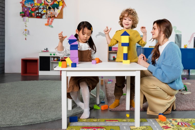 Duas crianças, uma menina e um menino, brincam com peças em cima de uma mesa. Uma mulher está agachada enquanto os observam. Fim da descrição.