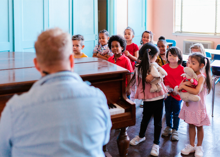 Grupo de crianças da educação infantil de diferentes raças observam professor sentado em frente a um piano. Fim da descrição.