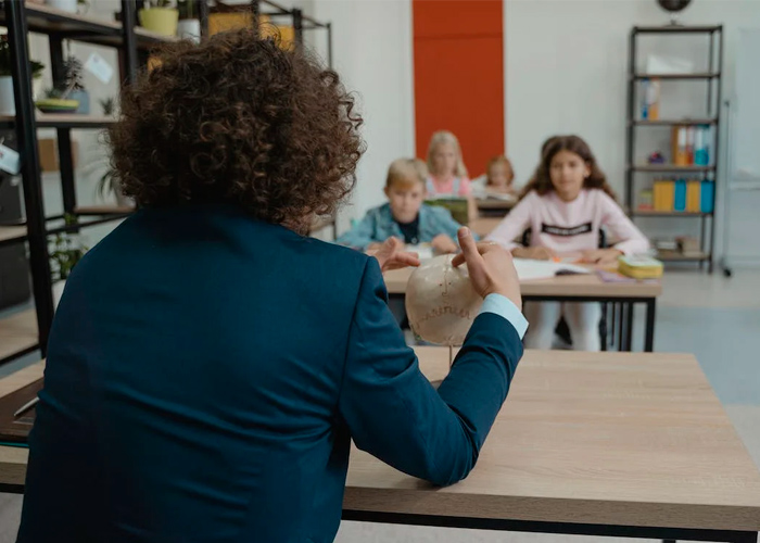 Em sala de aula, com estudantes ao fundo, educador aponta para réplica de crânio que está sobre sua mesa. Fim da descrição.