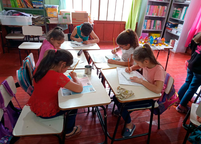  Em sala de aula, cinco estudantes pequenos estão sentados em grupo fazendo pinturas com tinta. Fim da descrição.