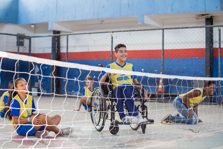 Cinco estudantes estão em quadra de esportes enquanto jogam vôlei sentado. Um menino está em cadeira de rodas. Fim da descrição.