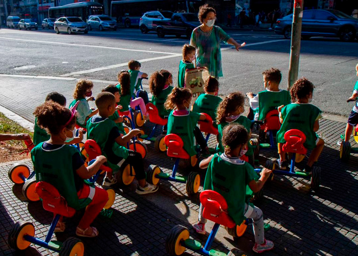 Grupo de crianças pequenas está em calçada com educadora à frente. Todas usam coletes verdes e estão em motocas. Fim da descrição.