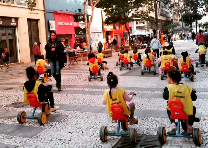 Em calçadão no centro de São Paulo, crianças com coletes amarelos pedalam motocas. Fim da descrição.