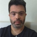 João Vitor posa para foto usando uma camiseta preta e uma corrente no pescoço, com pingente prata em formato de cruz. Ele é um homem branco na faixa etária de 30 anos, com barba, bigode e cabelo grisalhos. Fim da descrição.