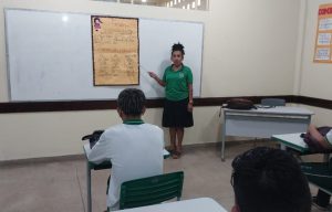 A professora Etelvina de Lira Gomes está em uma sala de aula, em pé, em frente à lousa. Ela é uma mulher de pele morena, veste uma camisa verde e uma saia preta, e está com os cabelos amarrados. À sua frente, estudantes sentados em suas carteiras prestam atenção à aula. 