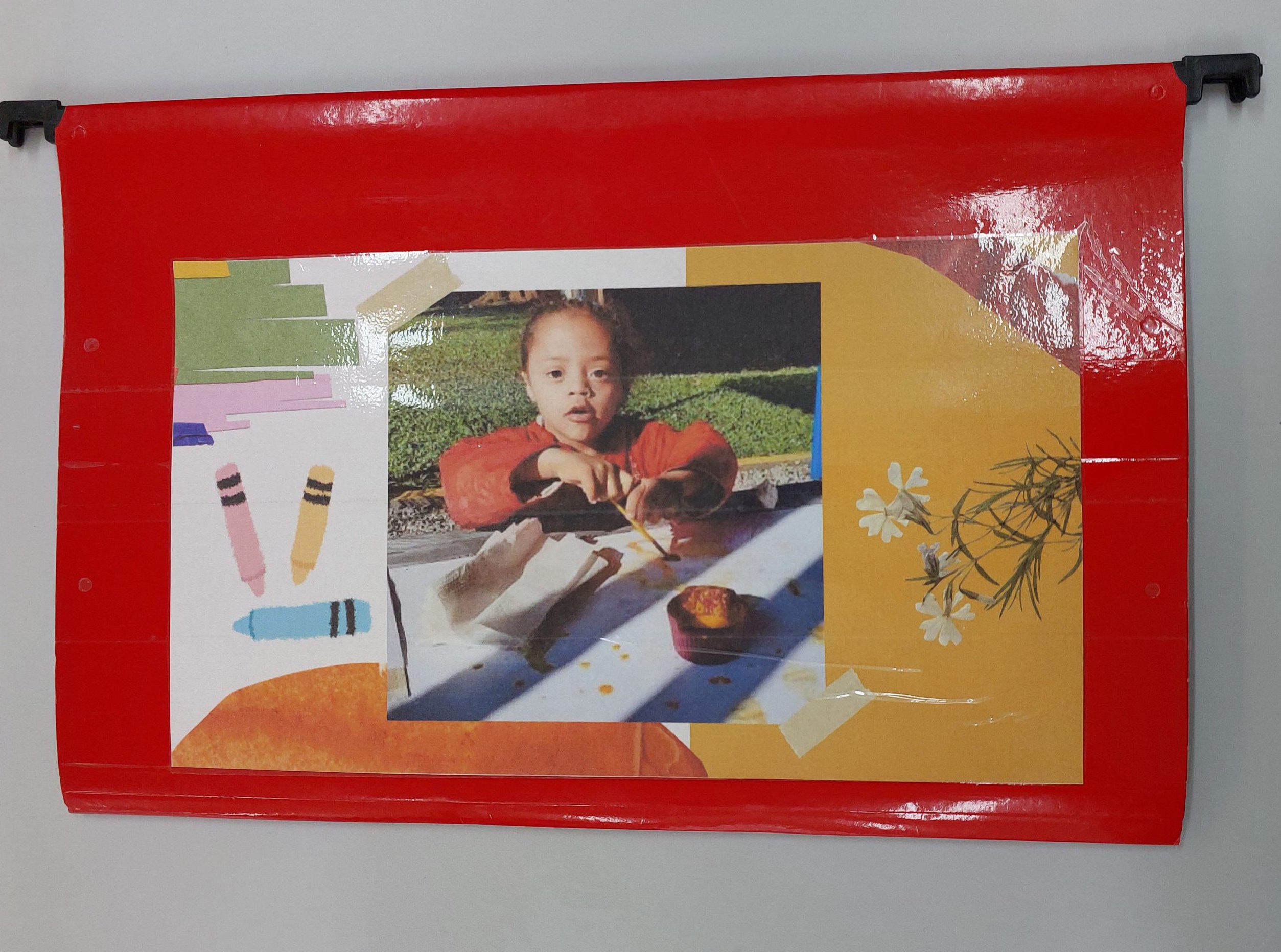 Pasta vermelha com foto de uma menina que está com pincel pintando em um papel sobre a mesa. Ao lado, o desenho de três lápis coloridos.