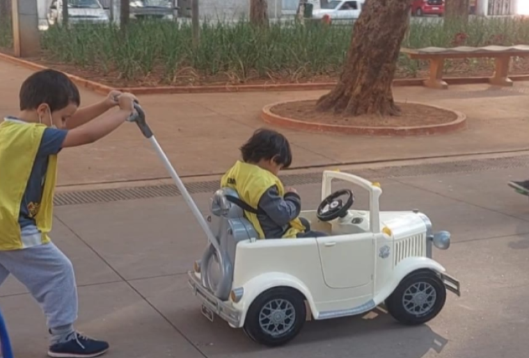 Um menino entre 4 e 5 anos de idade empurra um carrinho adaptado onde está outro menino. Eles passeiam em uma praça. Fim da descrição. 