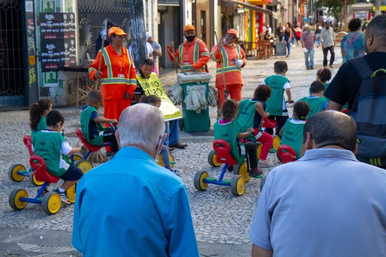 Um grupo de crianças entre 4 e 5 anos, vestindo coletes de identificação na cor verde, pedala pelo centro de São Paulo em motocas e interagem com os garis que trabalham na rua. Fim da descrição. 