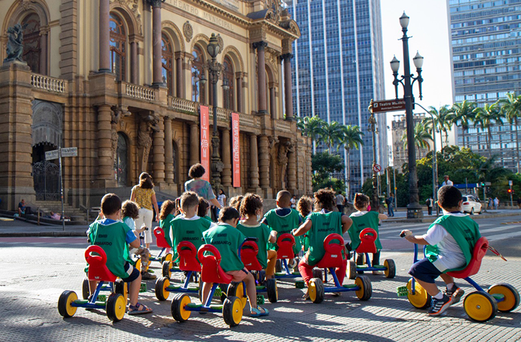 Um grupo de crianças entre 4 e 5 anos, vestindo coletes de identificação na cor verde, pedalam em motocas em frente ao Theatro Municipal de São Paulo durante um dia ensolarado, com duas professoras à frente. Fim da descrição.