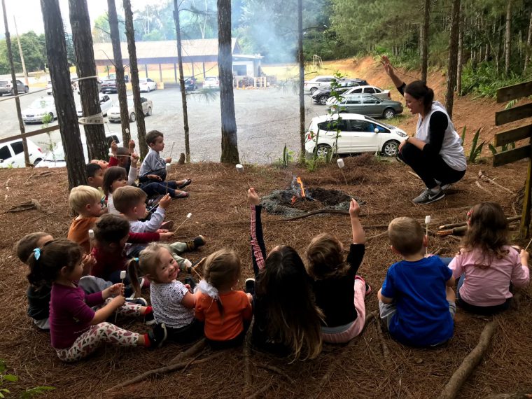 Uma professora e 16 crianças estão na área externa da escola, sentadas no chão, formando um semicírculo ao redor de uma fogueira. Eles seguram palitos com marshmallow na ponta. Fim da descrição