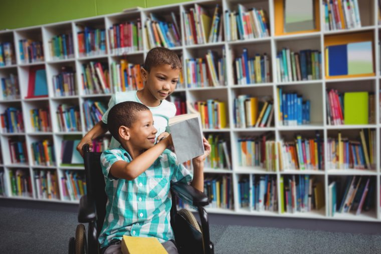 Dois garotos em frente a uma estante de livros: um está em cadeira de rodas e mostra um livro para o outro, que está atrás. Fim da descrição